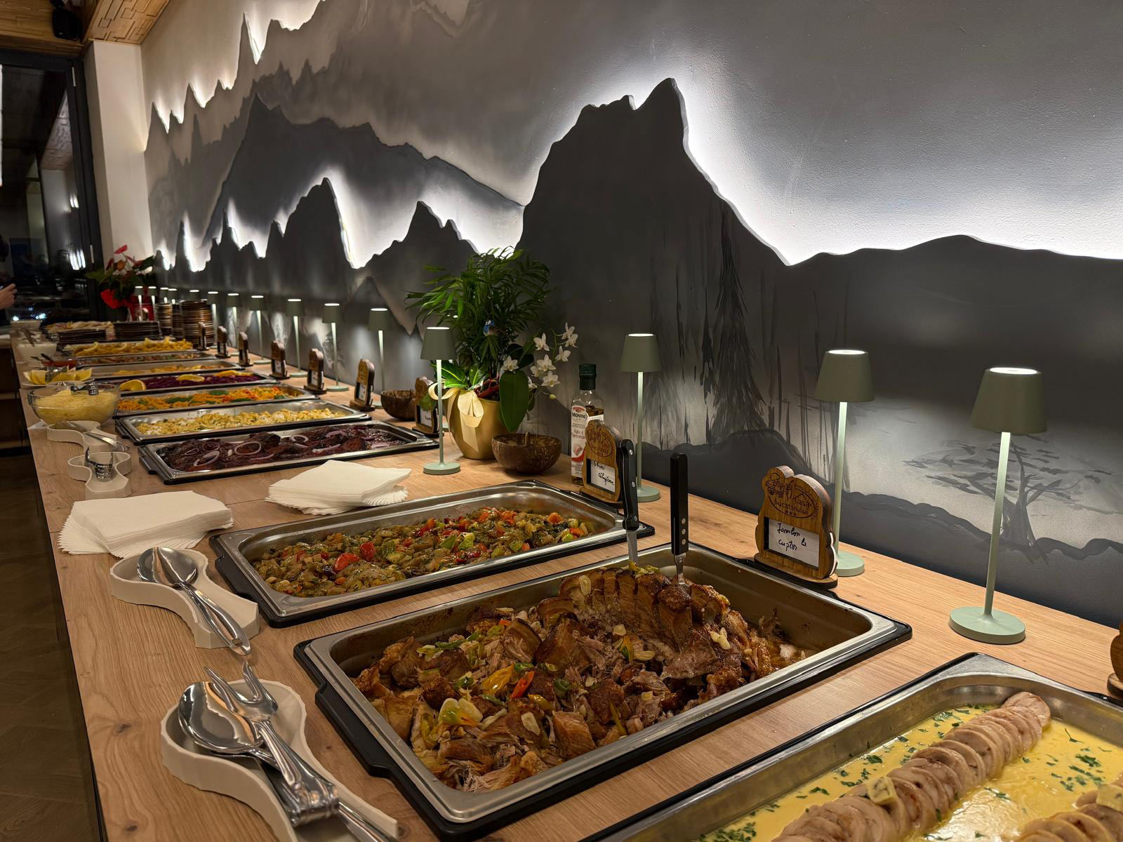 Buffet line with many hot dishes in metal trays along a wooden counter, mountain mural on the wall behind.