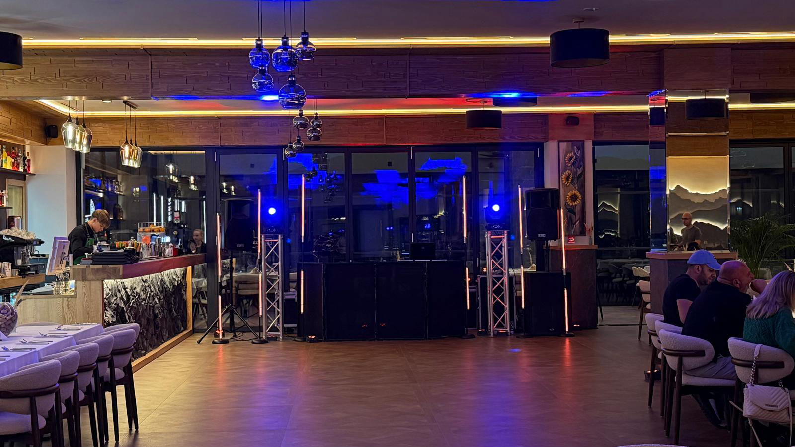 Bar area with a live music setup: DJ booth and speakers center stage, blue lights overhead and diners at tables along the sides.