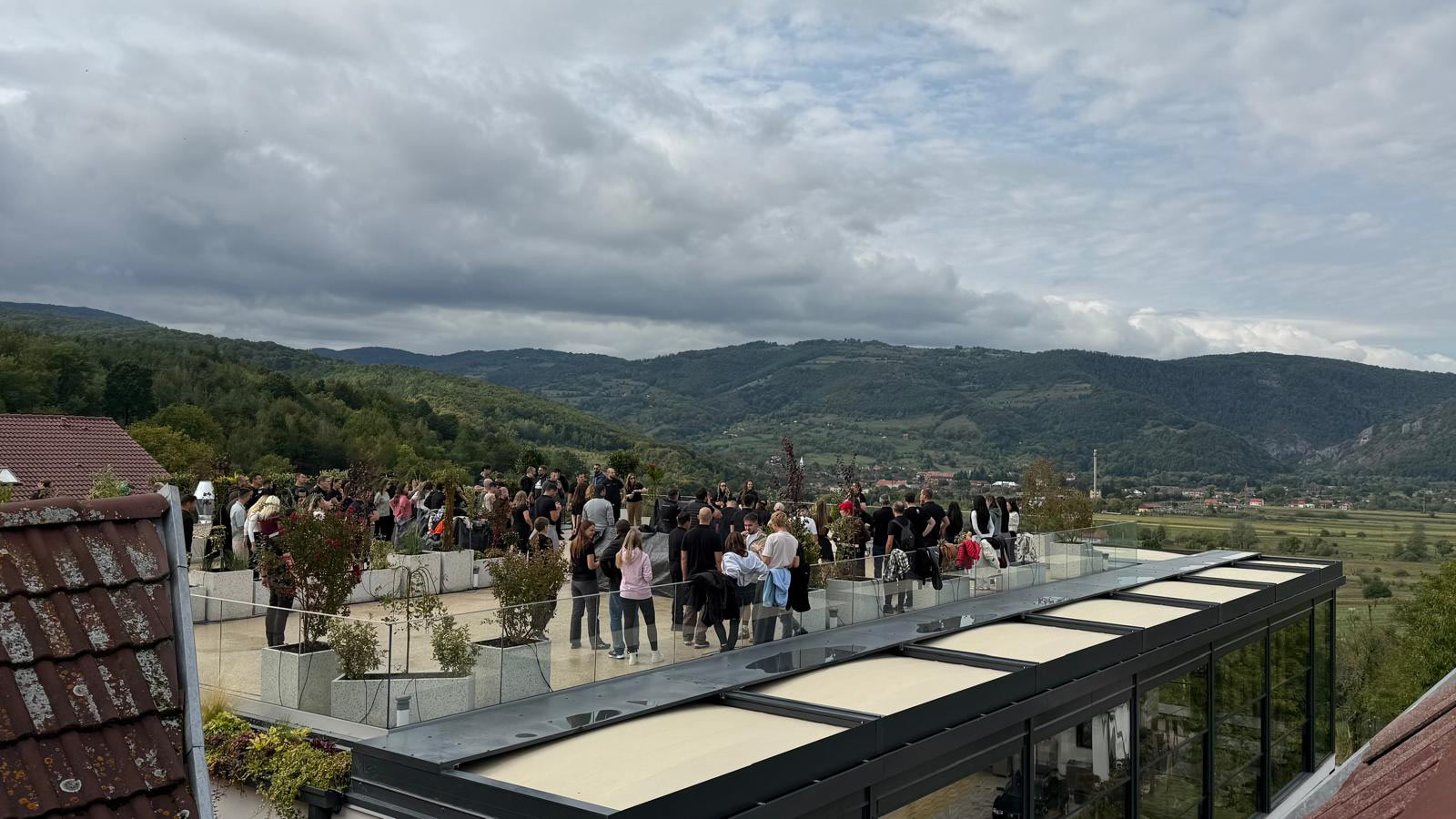 Group of people gathered on a modern rooftop terrace with glass railing and planters, overlooking green hills and distant mountains under a cloudy sky.