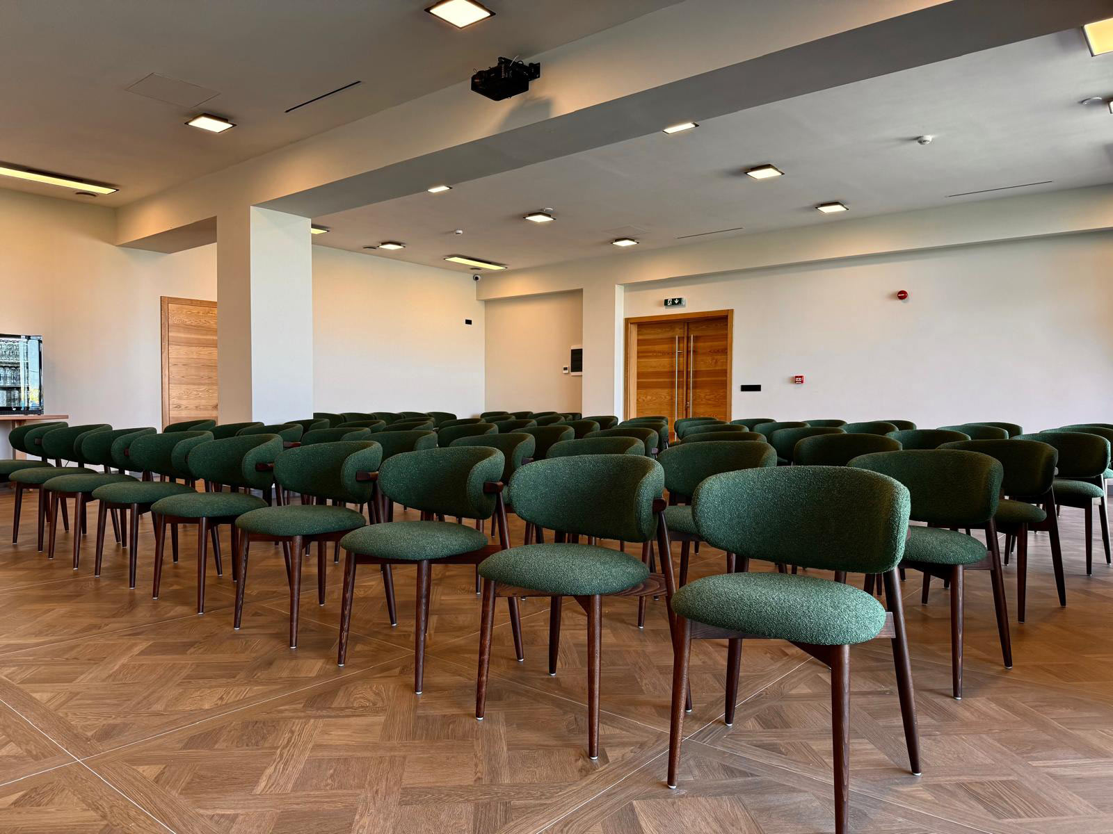 Empty conference room with rows of green upholstered chairs on a wood floor and a projector mounted to the ceiling.
