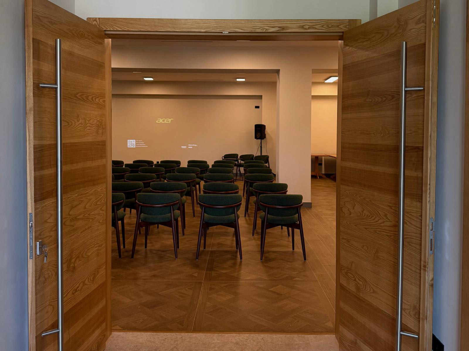 Double wooden doors open to a small conference room with green upholstered chairs arranged in rows facing a wall with an Acer logo on the projector screen area.