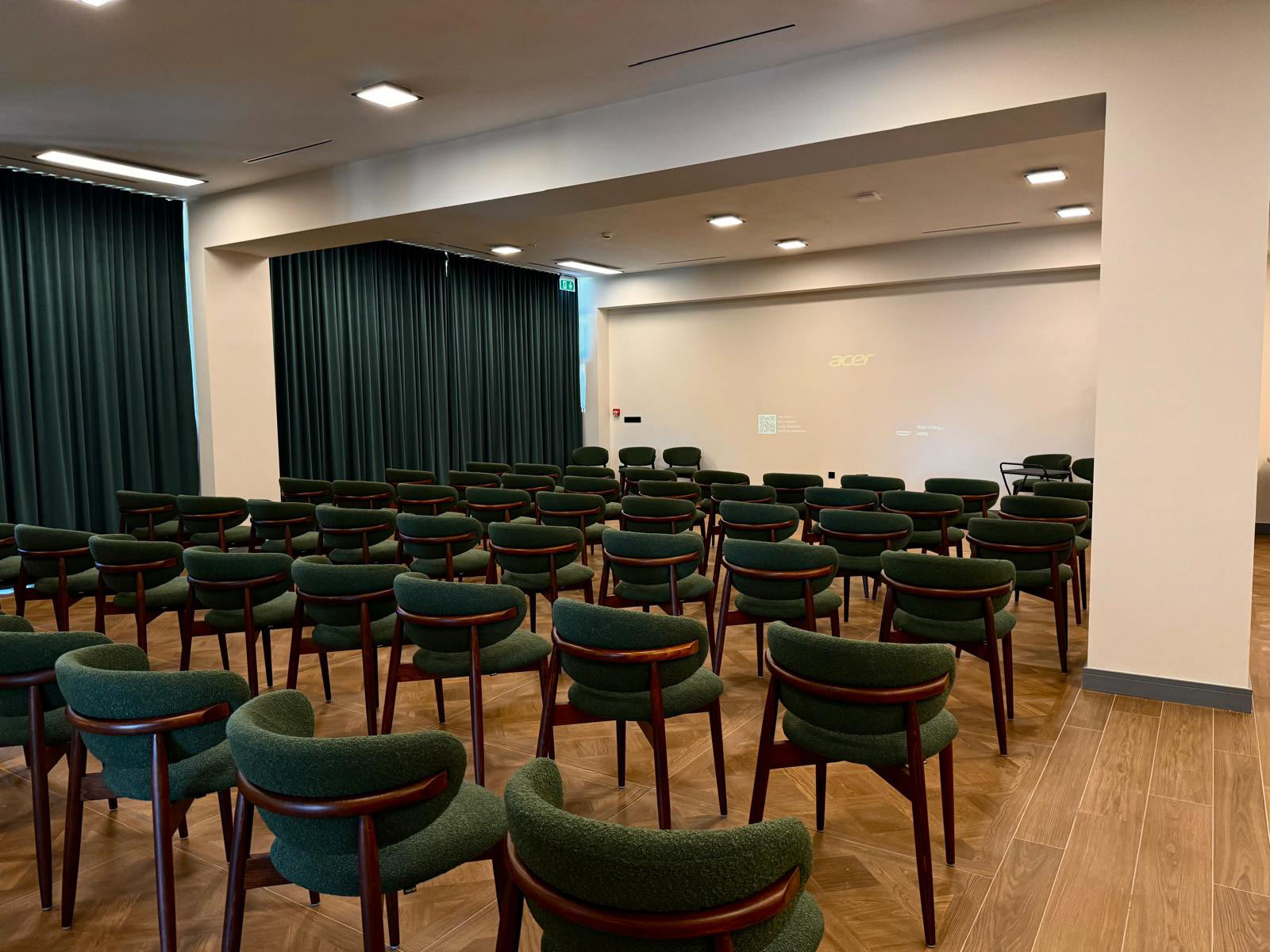Rows of green upholstered chairs in a small lecture hall with dark green curtains and a white wall showing an Acer logo projector.