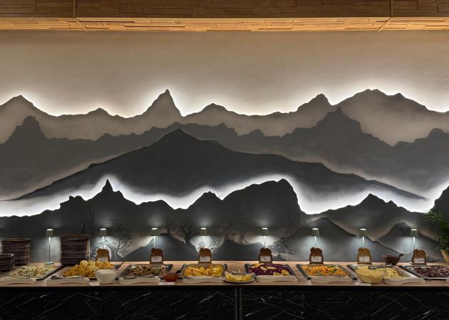 Buffet counter with a variety of dishes and warm lighting, set against a backlit mountain landscape mural on the wall.
